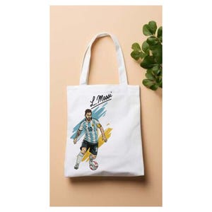 May include: A white tote bag featuring a graphic illustration of a soccer player in a light blue and white striped jersey with the number 10, dribbling a soccer ball. The design includes blue and yellow brush strokes and the text L. Messi.