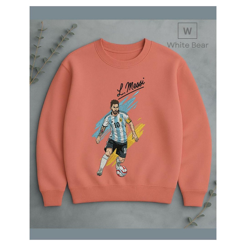 May include: A coral-colored crewneck sweatshirt featuring a graphic illustration of a soccer player in a light blue and white striped jersey with the number 10, dribbling a soccer ball, with the text L. Messi and White Bear logo.