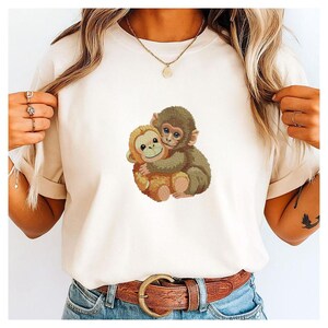 May include: Cream-colored t-shirt featuring an embroidered design of two hugging monkeys. One monkey is light brown, the other is dark brown. The shirt has short sleeves and a crew neck.