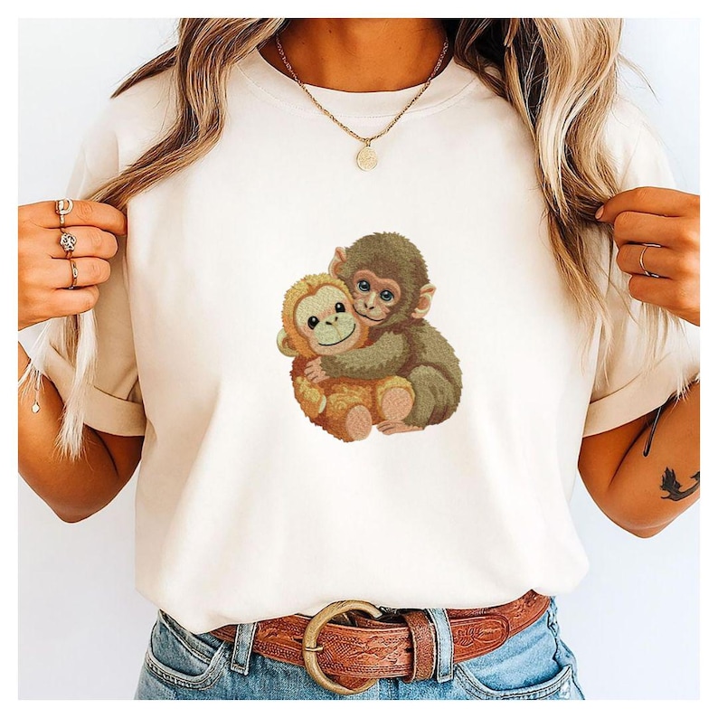 May include: Cream-colored t-shirt featuring an embroidered design of two hugging monkeys. One monkey is light brown, the other is dark brown. The shirt has short sleeves and a crew neck.
