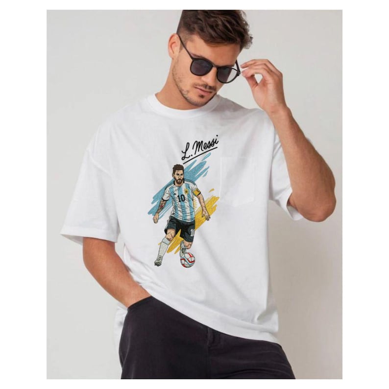 May include: A white short-sleeved t-shirt featuring a graphic illustration of a soccer player in a light blue and white striped jersey with the number 10, dribbling a soccer ball. The text L. Messi is printed above the figure in black script.