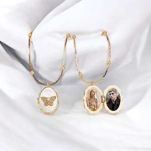 Puede incluir: Dos medallones ovalados dorados con cadenas. Uno presenta un diseño de mariposa sobre fondo blanco. El otro está abierto, revelando dos fotografías. Los medallones se exhiben sobre una superficie blanca.