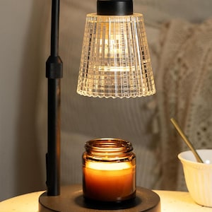 May include: A black candle warmer lamp with a clear, textured glass shade and a small jar candle. The lamp has a curved arm and a round base. The candle inside the jar is lit, casting a warm glow.
