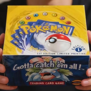 Puede incluir: Una caja de juego de cartas coleccionables Pokémon amarilla y azul. La caja presenta el logotipo de Pokémon, una Pokéball y el texto "Gotta catch 'em all!" y "1st Edition Limited Printing". La caja está sostenida por una persona.