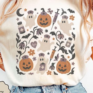 May include: An ivory t-shirt featuring a graphic print of ghosts wearing nurse caps, jack-o'-lanterns with nurse caps, syringes, medicine bottles, bats, flowers, a crescent moon, and a heart rhythm line in muted orange, brown, and dark gray tones.
