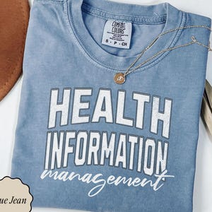 May include: A blue jean colored t-shirt with the words "HEALTH INFORMATION MANAGEMENT" in white block letters. The shirt has a crew neck and short sleeves. A gold necklace with a pendant is draped across the shirt.