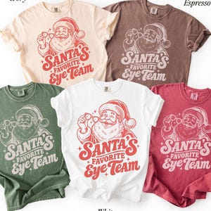 May include: Five short-sleeved t-shirts in ivory, espresso, moss, white, and crimson. Each shirt features a graphic of Santa Claus holding eyeglasses with the text "Santa's Favorite Eye Team" printed in a bold, stylized font with small star accents.