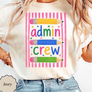 May include: An ivory t-shirt featuring a graphic with pink vertical stripes, three colored pencils in yellow, blue, and green, and the text "admin crew" in multicolored letters, accented by small colorful stars throughout the design.