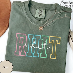 May include: A moss green short-sleeve t-shirt with a crew neckline. The front features the text RHIT in large, colorful block letters with a pink R, blue H, green I, and yellow T, overlaid with the word rhit in white cursive script.