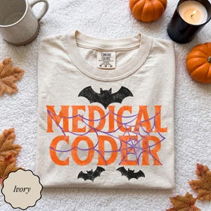 May include: An ivory t-shirt featuring the text MEDICAL CODER in orange block letters, overlaid with a purple spiderweb graphic. The design includes a black bat above the text and two smaller black bats below, with a Comfort Colors label.