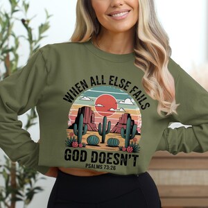 Puede incluir: Camiseta de manga larga verde oliva con un gráfico de paisaje desértico. El diseño presenta un sol rojo, cactus y el texto "WHEN ALL ELSE FAILS GOD DOESN'T" con "PSALMS 73:26" debajo. La camiseta es de estilo corto.