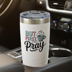 May include: A white travel mug with a silver rim and lid, featuring the text "BUT FIRST. PRAY" in black script and colorful block letters. A teal coffee cup graphic with a heart is also displayed. The mug is in a car's cup holder.