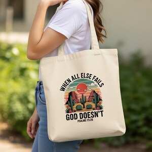 May include: Cream tote bag with a desert scene featuring cacti, mountains, and a sunset. The text reads "WHEN ALL ELSE FAILS GOD DOESN'T" with Psalms 73:26 below. The bag has two handles for carrying.