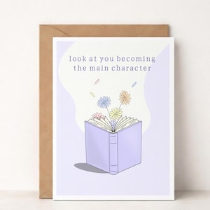 Op de afbeelding: Een wenskaart met een illustratie van een lichtpaars boek, met bloemen en de tekst "look at you becoming the main character". De kaart staat op een witte achtergrond met een bruine envelop.