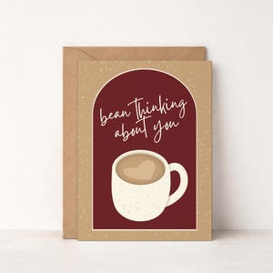 May include: A greeting card with a coffee-themed design. The card features a white mug with a heart-shaped coffee design, set against a burgundy background. The text "bean thinking about you" is written in cursive. The card is paired with a brown envelope.