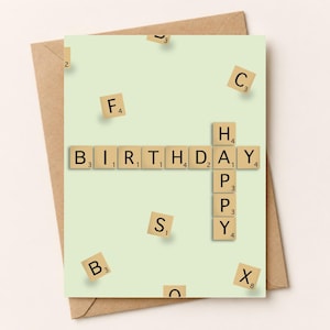 May include: A light green greeting card featuring tan letter tiles arranged to spell BIRTHDAY and HAPPY in a crossword style. Additional scattered letter tiles including F, C, S, B, and X are distributed across the card surface.