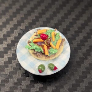 May include: Miniature food item on a small plate. The dish features a small, round base topped with green lettuce, orange cheese, and a red cherry tomato. Two green olives with red accents are on the plate. The plate has a blue and white checkered pattern.