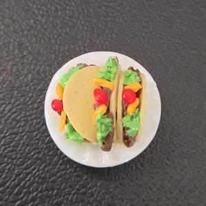 May include: A miniature food replica featuring two tacos served on a small white scalloped plate. Each taco consists of a tan shell filled with brown meat, bright green lettuce, orange shredded cheese, and a single small red tomato piece.