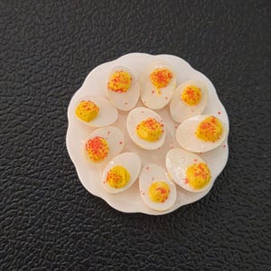 May include: A miniature white scalloped plate holding nine halved deviled eggs. Each egg white half features a bright yellow yolk center topped with a dusting of red paprika-like seasoning. The food items are arranged in a circular pattern.