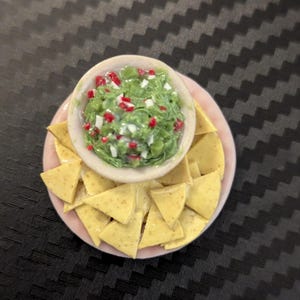 May include: Miniature food item featuring a bowl of guacamole with red and white accents, surrounded by yellow tortilla chips on a pink plate. The food is arranged on a black surface.