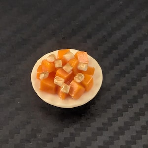 May include: A miniature dish featuring orange diced food, possibly vegetables, topped with small, light-colored cubes. The food is arranged on a small, white, circular plate. The background is a dark, textured surface.