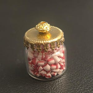 May include: A miniature clear glass jar filled with small, cylindrical white and red striped candy pieces. The jar is topped with a gold-colored metal lid featuring a decorative scalloped edge and a small, textured knob handle on top.