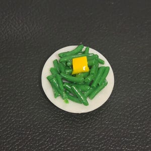 May include: A miniature white plate topped with a pile of glossy green beans and a single square piece of bright yellow butter resting in the center. The food items are rendered with a smooth, shiny finish on a small, round, white dish.