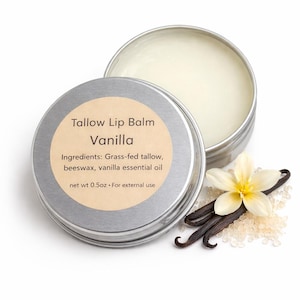 May include: A silver tin of vanilla tallow lip balm, with the lid slightly ajar. The label reads "Tallow Lip Balm Vanilla." Ingredients include grass-fed tallow, beeswax, and vanilla essential oil. The net weight is 0.5 oz. Vanilla beans and a flower are next to the tin.