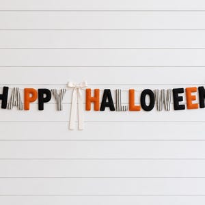 Puede incluir: Una guirnalda decorativa que deletrea HAPPY HALLOWEEN con letras de tela individuales en negro, naranja y patrones de rayas grises y blancas. Las letras están ensartadas en un cordel y decoradas con tres lazos de tela color crema.