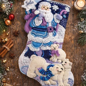 Könnte beinhalten: Handgefertigter Weihnachtsstrumpf mit Weihnachtsmann und Eisbären. Der Strumpf ist hauptsächlich blau, weiß und lila, mit Pailletten- und Filzdetails. Der Name "Julia" ist auf einem wolkenförmigen Anhänger gestickt. Verziert mit Schneeflocken und festlichen Elementen.