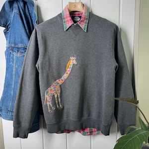 May include: A gray sweatshirt with a colorful giraffe applique. The sweatshirt has a collared shirt underneath with a pink and green plaid pattern. A denim jacket hangs in the background.
