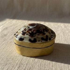 Peut inclure: Une petite boîte ovale avec couvercle, dotée d'un dessus en coquillage poli aux motifs naturels tachetés de brun et de crème. La base du récipient présente une finition dorée mate unie, créant un contraste marqué entre le couvercle en coquillage organique et la section inférieure métallique de l'objet.