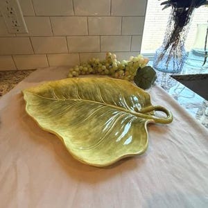 Op de afbeelding: Een grote, blad-vormige serveerschaal in groen- en geeltinten. De keramische schaal heeft gedetailleerde aderen en een gebogen handvat. De schaal is ongeveer 51 cm lang en 38 cm breed.