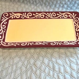 May include: Rectangular serving platter with a yellow centre and a burgundy border. The border features a white, stylised floral design. The platter is placed on a light blue quilted surface.