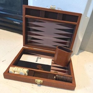 May include: A brown leather-covered backgammon board game set featuring a hinged case with a gold-tone metal latch. The interior displays a playing surface with dark and light brown triangular points, white and dark checkers, and a leather cup.