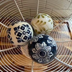 May include: Three decorative ceramic spheres with intricate patterns sit within a white wire basket. One sphere has a blue and white floral design, another has green leaf patterns, and the third has a dark blue floral design.