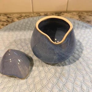 以下が含まれることがあります： 青いセラミック製のクリーマーと、それに合う蓋。クリーマーは、湾曲した注ぎ口と薄いタン色の縁を持つユニークな形状をしています。蓋も青色で、同様の形状をしています。クリーマーは模様のある表面に置かれています。