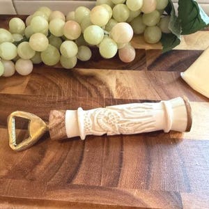 May include: A decorative bottle opener with a carved white handle and a gold-colored opener. The handle features a detailed relief design. The opener is lying on a wooden cutting board, with grapes and cheese in the background.