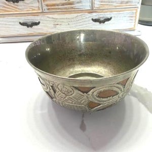 May include: A silver-colored metal bowl with an embossed design around the rim. The bowl has a copper-colored band and a decorative pattern of stylized figures. The bowl is sitting on a white surface.
