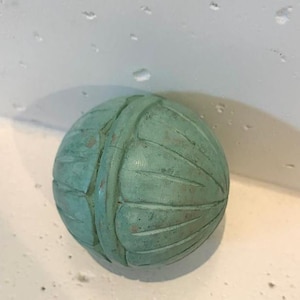 May include: A spherical object with a light green, weathered finish. The surface features carved, vertical, leaf-like segments radiating from a central band that encircles the sphere, creating a textured, segmented appearance across the entire item.