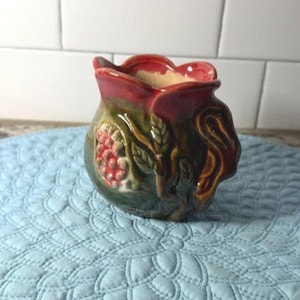 May include: A small ceramic pitcher featuring a scalloped rim with a glossy red finish. The body is dark green with a raised, textured relief of red berries and green leaves on the side. The handle is shaped like a curved, brown vine.
