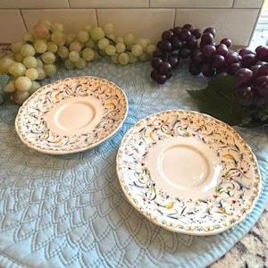 May include: Two round ceramic saucers featuring a cream-colored base with a decorative border of swirling blue, yellow, and green floral patterns. The edges of the saucers are scalloped, and the center of each piece is smooth and plain.