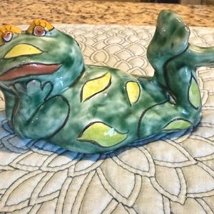 Könnte beinhalten: Keramik-Froschfigur in entspannter Pose. Der Frosch ist überwiegend grün mit gelben Flecken und Akzenten. Er hat ein skurriles Design mit einem lächelnden Ausdruck und liegt auf einer gemusterten Oberfläche.
