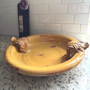 Puede incluir: Un cuenco grande y redondo de cerámica amarilla con asas ornamentadas en forma de hojas. El cuenco tiene un acabado envejecido con detalles marrones. En el fondo hay una tabla de cortar negra decorativa con una flor morada. El cuenco es probablemente para fines decorativos o de servicio.
