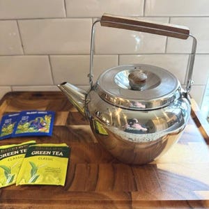 以下が含まれることがあります： 木製のハンドルと蓋のつまみが付いた、光沢のあるステンレス製のティーポット。木製のまな板の上に置かれ、緑茶と安眠ティーのパックが並んでいます。ティーポットは周囲の環境を反射しています。