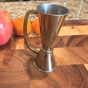 Könnte beinhalten: Ein Cocktail-Jigger aus Edelstahl mit Griff, neben einem roten Apfel und zwei Orangen. Der Jigger hat eine konische Form mit ausgestelltem Boden und einem Messbecher oben. Der Griff ist gebogen und an der Seite befestigt.