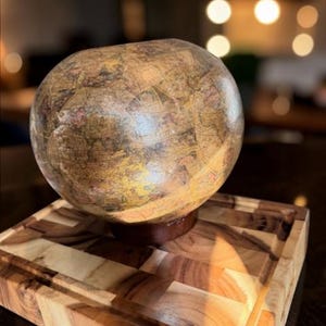 May include: A spherical, antique-style globe with a map design, resting on a wooden base. The globe is set on a wooden cutting board with a carved groove. The globe's colors include shades of brown, beige, and red.