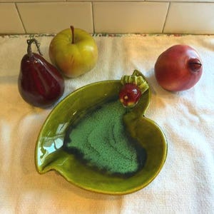 May include: A glossy, leaf-shaped ceramic dish in shades of lime green and dark teal, featuring a small decorative apple accent on the rim. The dish is surrounded by a dark red pear, a yellow-green apple, and a round, reddish-brown pomegranate.