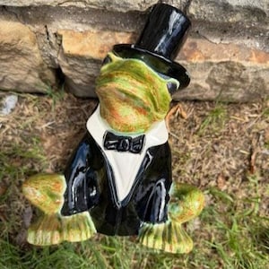May include: A ceramic frog figurine in a black tuxedo, bow tie, and top hat. The frog has a green and orange face and feet. The figurine is posed as if sitting.