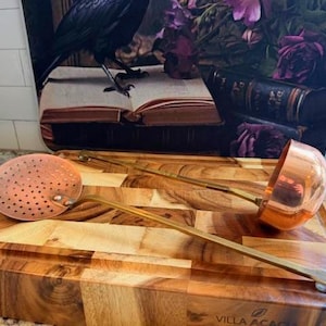 May include: A copper skimmer and ladle with brass handles rest on a wooden cutting board. The skimmer is round with small holes. The ladle has a rounded bowl. The cutting board has a geometric pattern. A decorative background is visible.